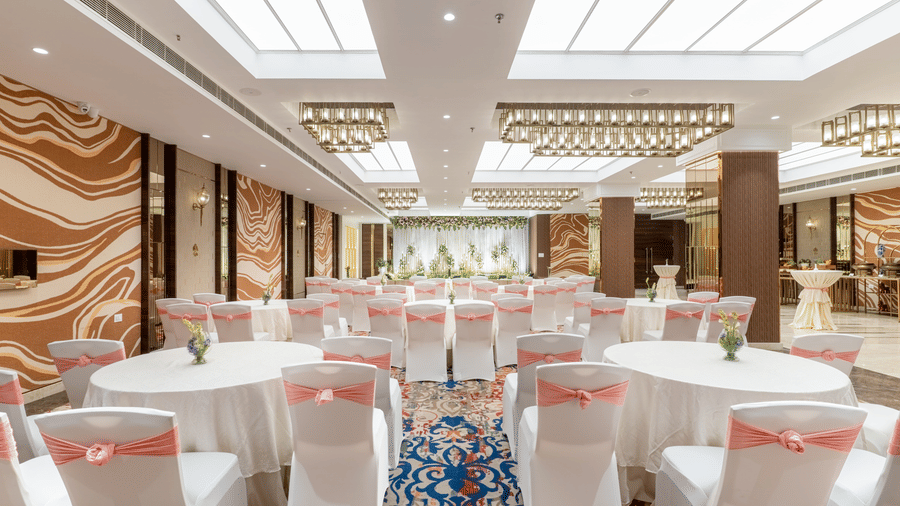 A banquet hall at Nirvana Sarovar Portico, Gorakhpur featuring round tables, covered chairs, and a decorated background stage.