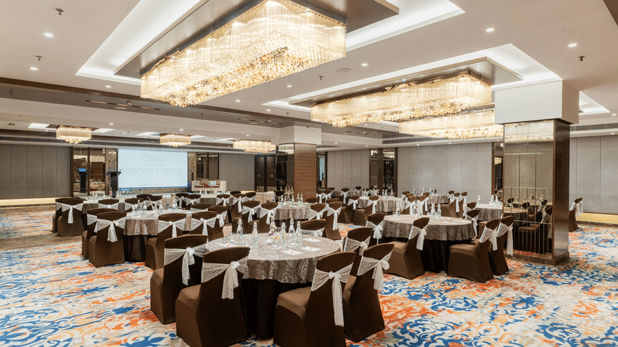 A banquet hall at Nirvana Sarovar Portico, Gorakhpur featuring round tables, dressed chairs, chandeliers, and a projection screen. 