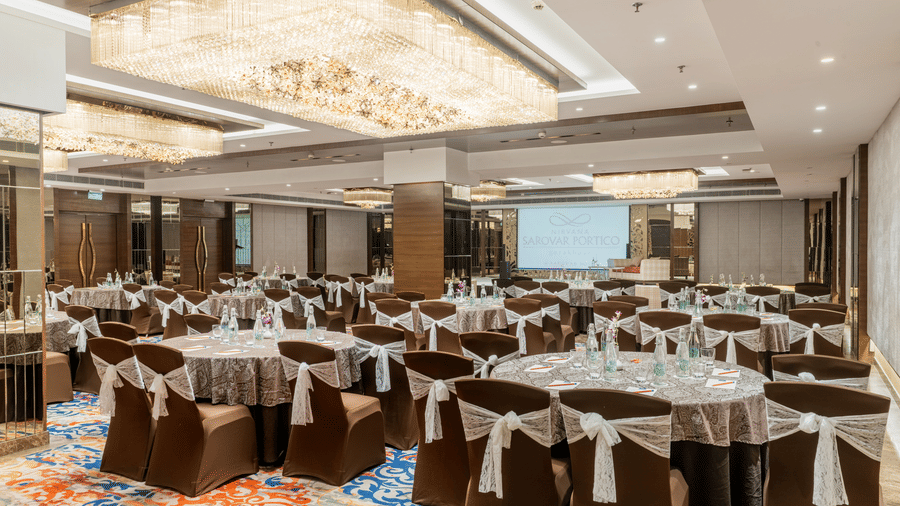 A banquet hall at Nirvana Sarovar Portico, Gorakhpur featuring round tables, dressed chairs, and patterned carpets.