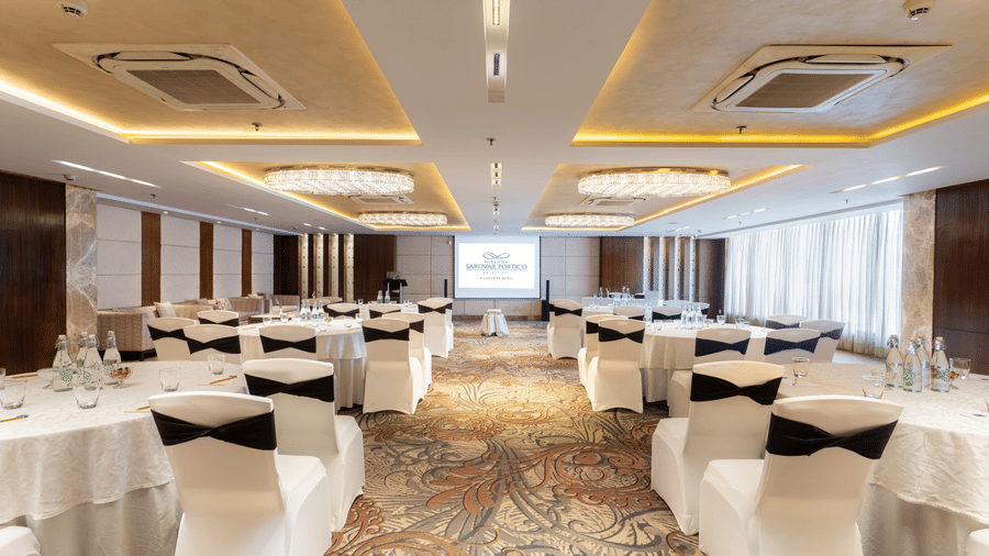 A conference setup at Nirvana Sarovar Portico, Gorakhpur featuring round tables, covered chairs, and a projection screen.