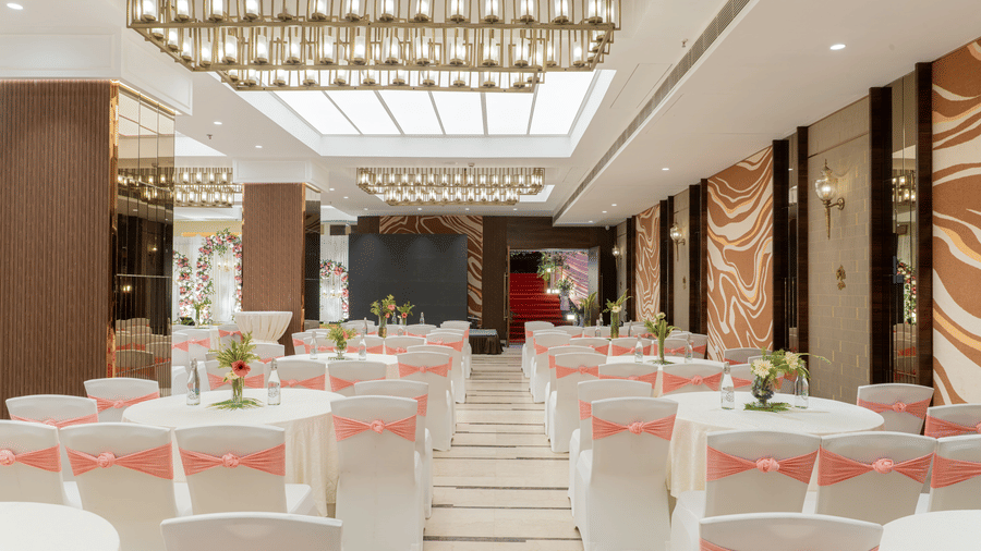 A function hall at Nirvana Sarovar Portico, Gorakhpur featuring round tables, dressed chairs, chandeliers, and decorative wall panels. 