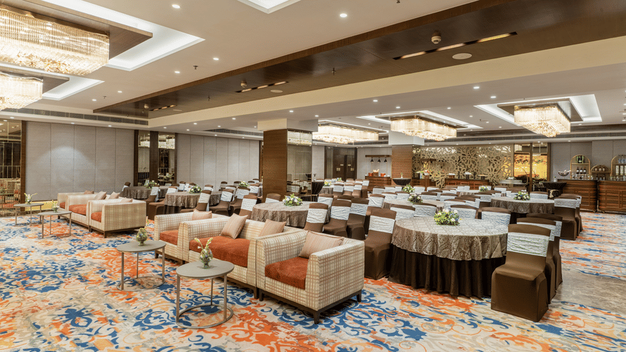 A large hall at Nirvana Sarovar Portico, Gorakhpur featuring plush sofa seating, round dining tables, and chandelier lighting.