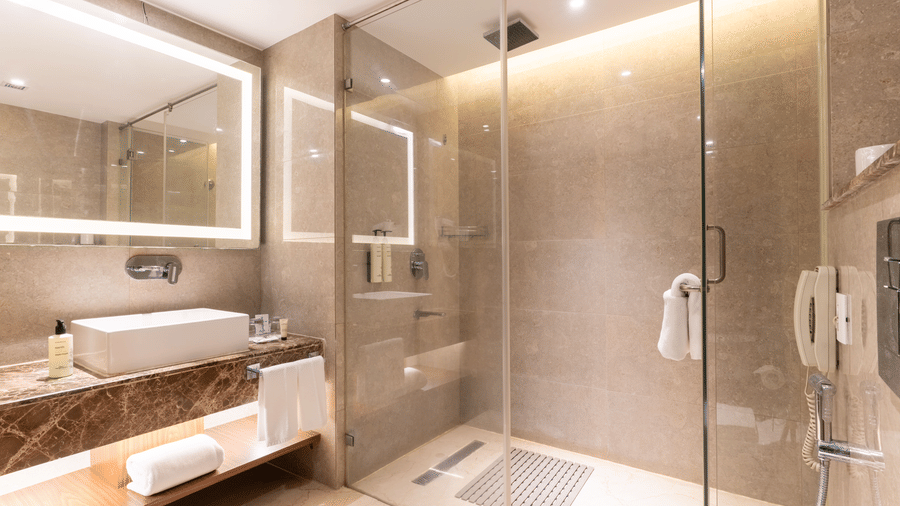 A bathroom at Nirvana Sarovar Portico, Gorakhpur featuring a walk in glass shower, a washbasin, a large illuminated mirror, and bath towels.  