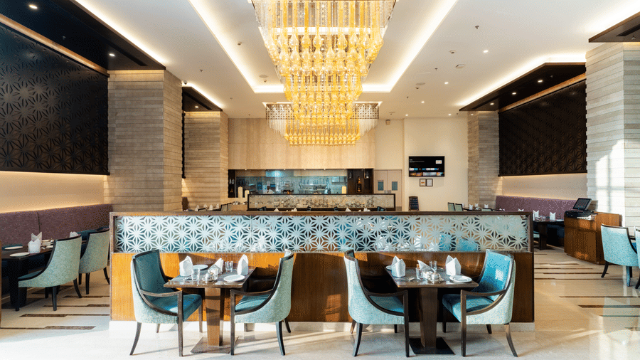 A dining area at Nirvana Sarovar Portico, Gorakhpur featuring dining tables, padded chairs, and chandeliers.