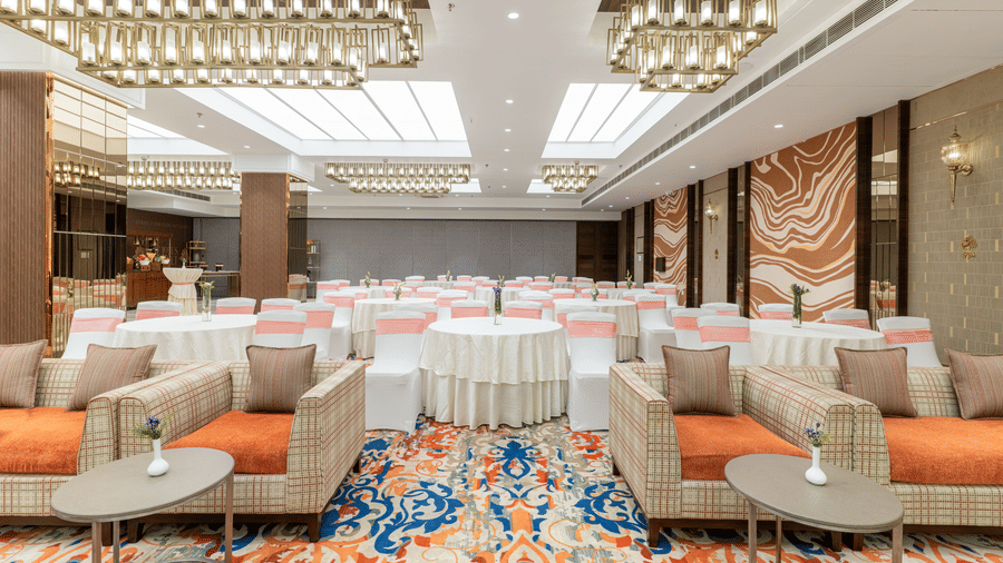 An event hall at Nirvana Sarovar Portico, Gorakhpur featuring upholstered sofas, small coffee tables, and round dining tables with chairs. 