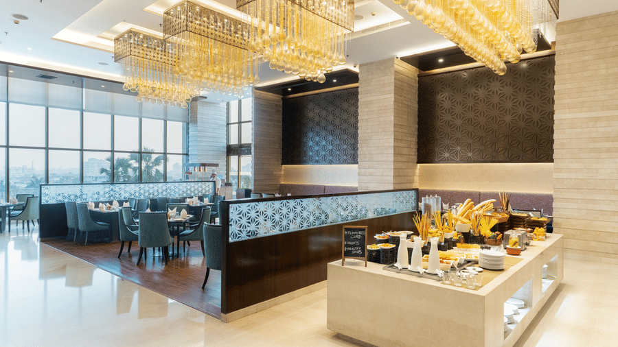 A restaurant dining room at Nirvana Sarovar Portico, Gorakhpur featuring a buffet station, dining tables, chairs, and large windows. 
