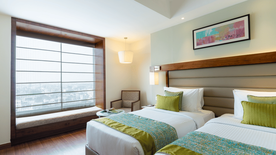 A hotel bedroom at Nirvana Sarovar Portico, Gorakhpur featuring two twin beds, a window bench with city views, and modern artwork.