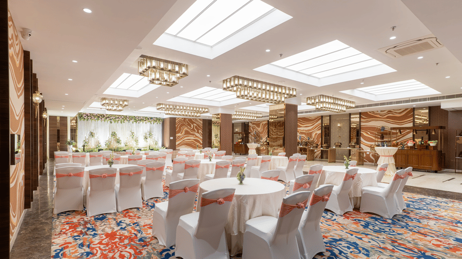 A decorated hall at Nirvana Sarovar Portico, Gorakhpur featuring round tables, covered chairs, and a decorated stage.