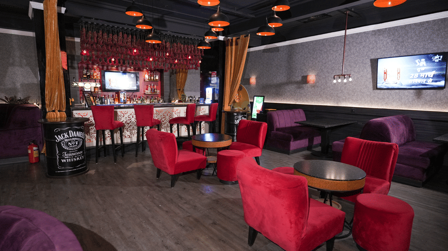 Wide view of a lounge at Park Plaza Ludhiana featuring red sofas and chairs dark ceiling with orange pendant lights a bar counter in the background and a wall-mounted TV.