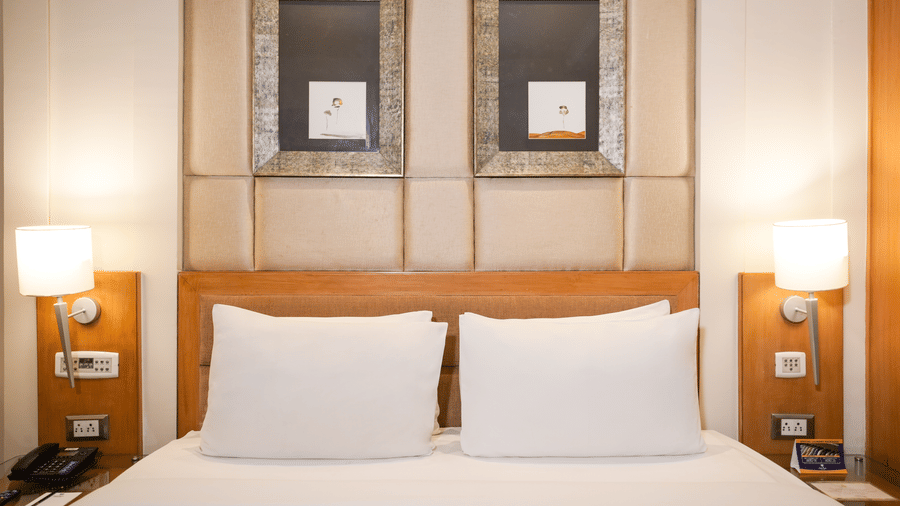 Close up view of a bed at Park Plaza Ludhiana featuring white pillows and bedding a wooden headboard with framed panels and warm bedside lighting on both sides.