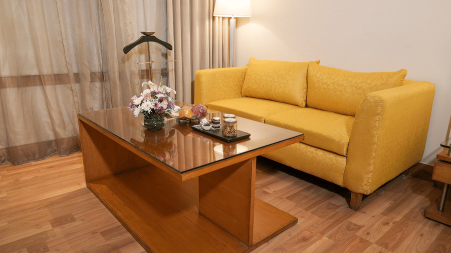A lounge area at Park Plaza Ludhiana featuring a yellow sofa a wooden coffee table with flowers sheer curtains a floor lamp and warm wooden flooring in a bright setting.