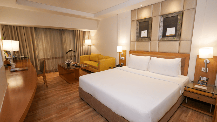 A room at Park Plaza Ludhiana featuring a king bed with white bedding wooden furniture a yellow sofa warm lighting and a work desk area in a modern setting.