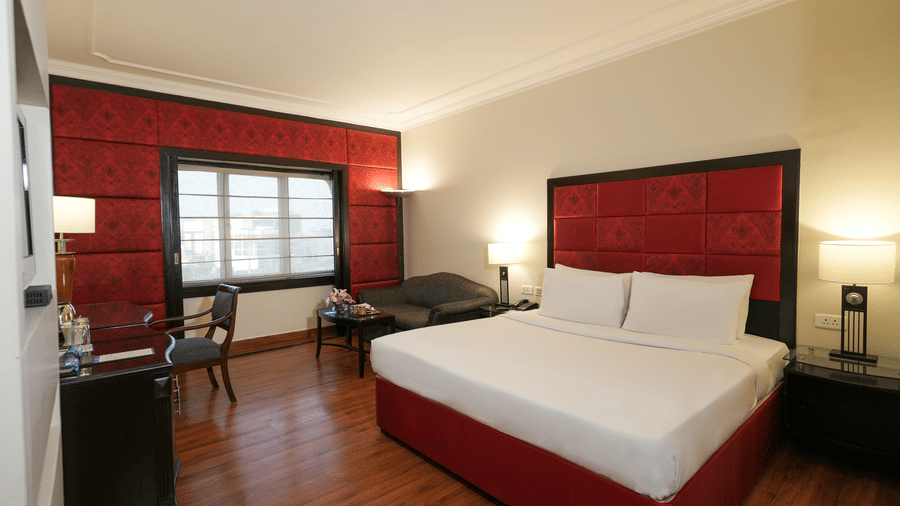 A room at Park Plaza Ludhiana featuring a king bed with white bedding and bold red headboard panel dark wooden flooring bedside lamps and a seating area near the window.