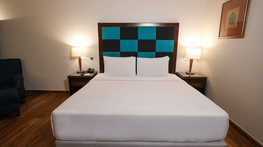 A room at Park Plaza Ludhiana featuring a king bed with white bedding and teal checkered headboard wooden flooring bedside tables with lamps and warm ambient lighting.