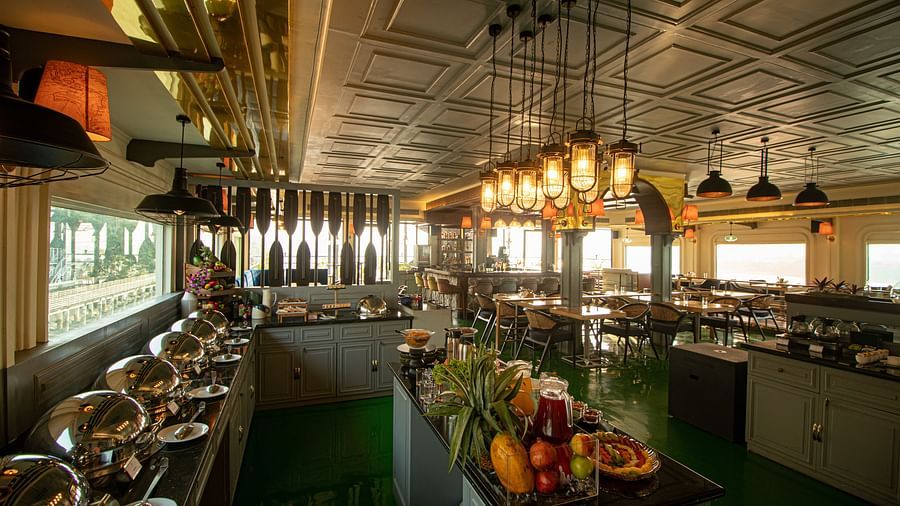 An elegant hotel restaurant buffet with chafing dishes, a fruit display, and lantern-style pendant lighting, with views of the outdoors.