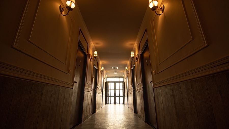 A dimly lit hotel corridor lined with wooden doors and wall-mounted lights, leading towards a lit exit.