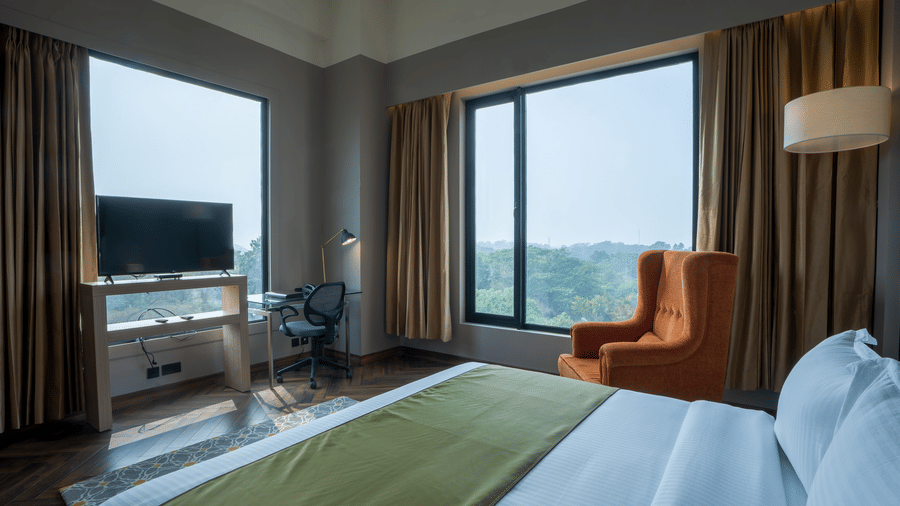 Bedroom with king bed, work desk, orange armchair and large window overlooking greenery at Polo Hotel, Agartala.