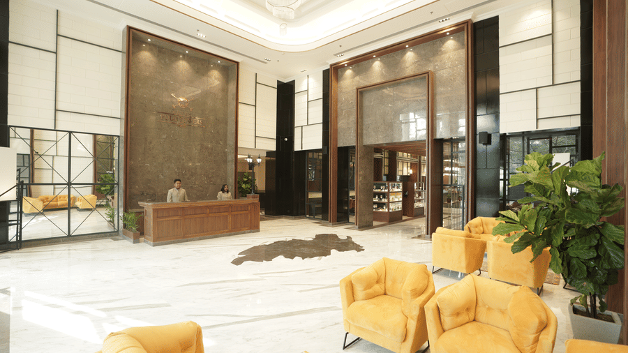 Reception lobby at Polo Hotel, Agartala with a wooden front desk, two staff members, mustard armchairs, marble floor and tall windows.