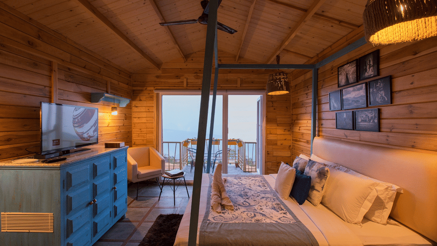 Wood cabin room at Polo Resort Cherrapunji with four-poster bed, blue dresser with TV, framed photo wall and a guest standing at balcony doors at dusk.