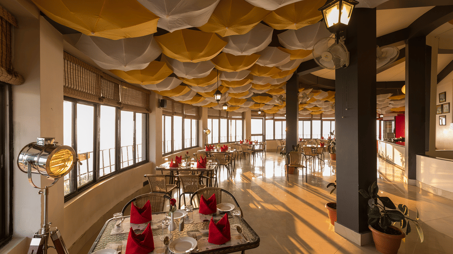 Polo Resort Cherrapunji restaurant interior with yellow and white umbrella ceiling, wicker chairs, red napkin tables and large valley-view windows.