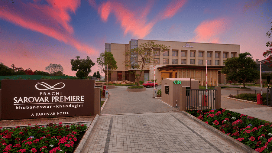 Entrance driveway at Prachi Sarovar Premiere, Bhubaneswar, with a hotel signpost, flowerbeds, and the main building beneath a twilight sky.
