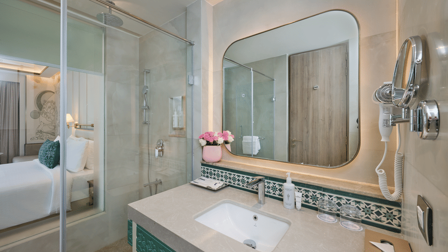 Bathroom at Prachi Sarovar Premiere, Bhubaneswar, with a stone washbasin vanity, a large mirror, a toilet, and a glass-enclosed shower area.