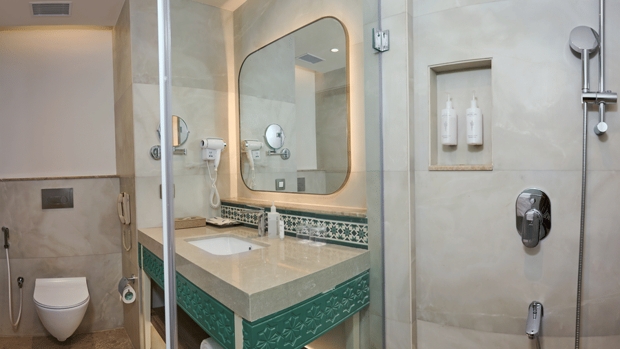 Bathroom at Prachi Sarovar Premiere, Bhubaneswar, with a stone washbasin vanity, a large mirror, a toilet, and a glass-enclosed shower area.