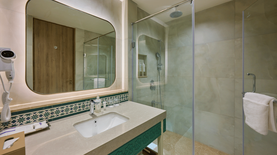 Vanity area at Prachi Sarovar Premiere, Bhubaneswar, displaying a washbasin with a mirror, toiletries, and an adjacent glass shower stall.