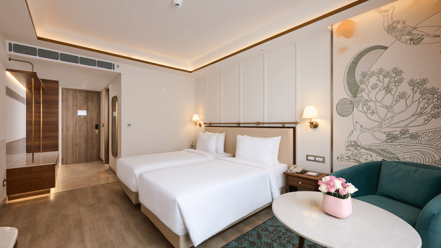 Twin bedroom at Prachi Sarovar Premiere, Bhubaneswar, with 2 single beds, a sofa, an oval coffee table, wall lamps, and a room entrance.