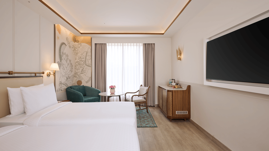 Deluxe Twin Room view at Prachi Sarovar Premiere, Bhubaneswar, with beds, a television, a wooden unit, an armchair, and a window with curtains.