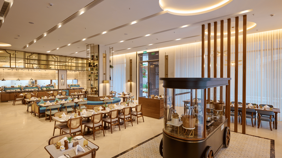 Flavours dining hall at Prachi Sarovar Premiere, Bhubaneswar, with long wooden tables, chairs, a decorative food cart, and ceiling lights.