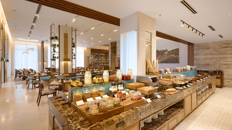Buffet setup at Prachi Sarovar Premiere, Bhubaneswar, showing a long stone counter with food items, dining tables, and overhead lighting fixtures.