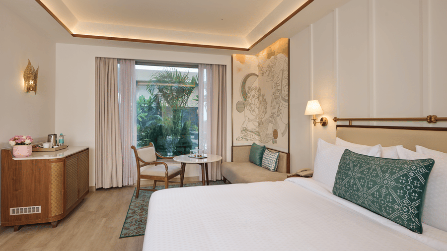 Premium room at Prachi Sarovar Premiere, Bhubaneswar, showing a bed, a wooden cabinet, a seating area by the glass door, and wall panels.