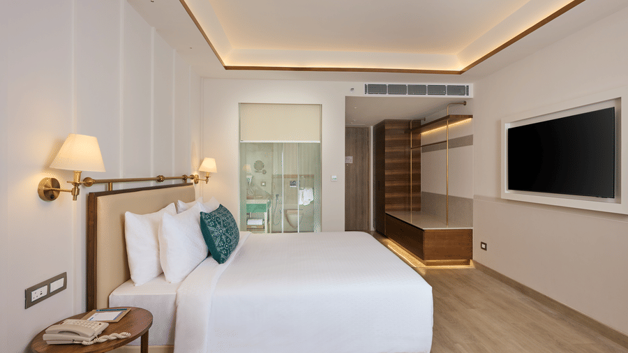 Premium Room at Prachi Sarovar Premiere, Bhubaneswar, featuring a bed with cushions, wall lamps, a wall-mounted television, and a doorway.