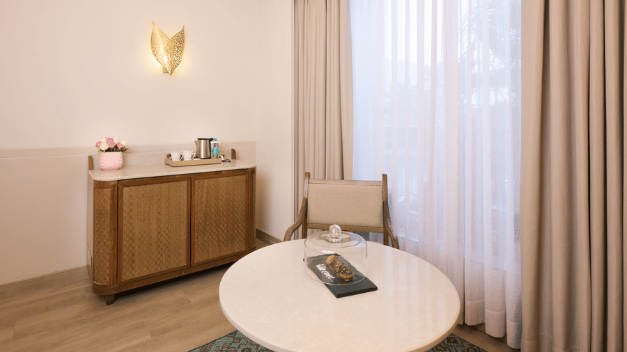 Seating area at Prachi Sarovar Premiere, Bhubaneswar, with a round table, a chair, a wooden cabinet, and curtains next to a window.