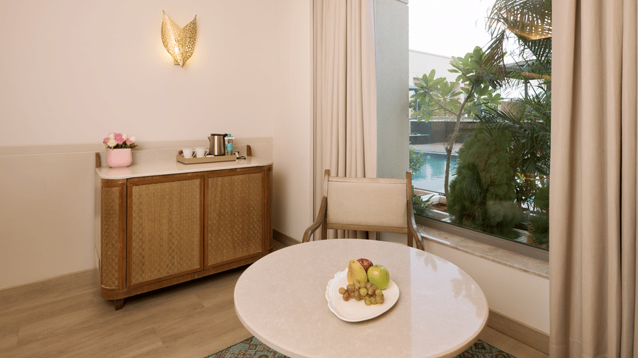 Seating area at Prachi Sarovar Premiere, Bhubaneswar, with a round table, a chair, a wooden cabinet, and curtains next to a window.