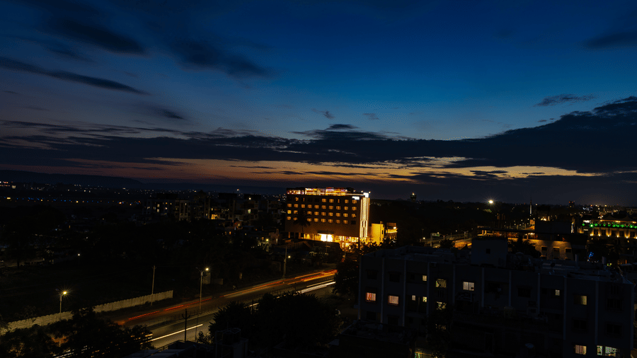 An aerial view of a city at dusk, showing street lights and buildings, with a prominent multi-storey building in the middle distance against a dark blue and orange sky