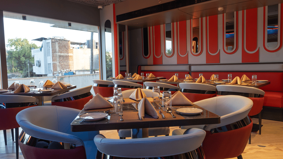 A restaurant interior with individual tables set with cutlery, featuring chairs, booth seating with red accents, and large windows | Pride Elite Chhatrapati Sambhajinagar