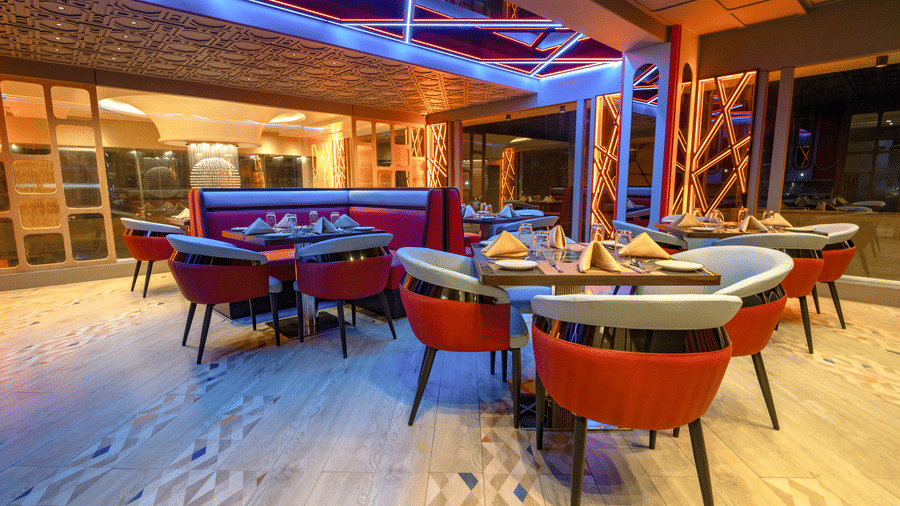 A dining area with tables set with chairs on a light-coloured floor, under a wooden roof structure, illuminated with blue lights at night | Pride Elite Chhatrapati Sambhajinagar