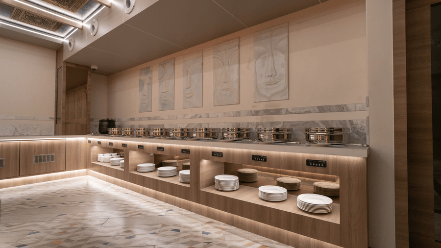 A brightly lit buffet serving station with multiple chafing dishes and plates, featuring wood and marble accents | Pride Elite Chhatrapati Sambhajinagar