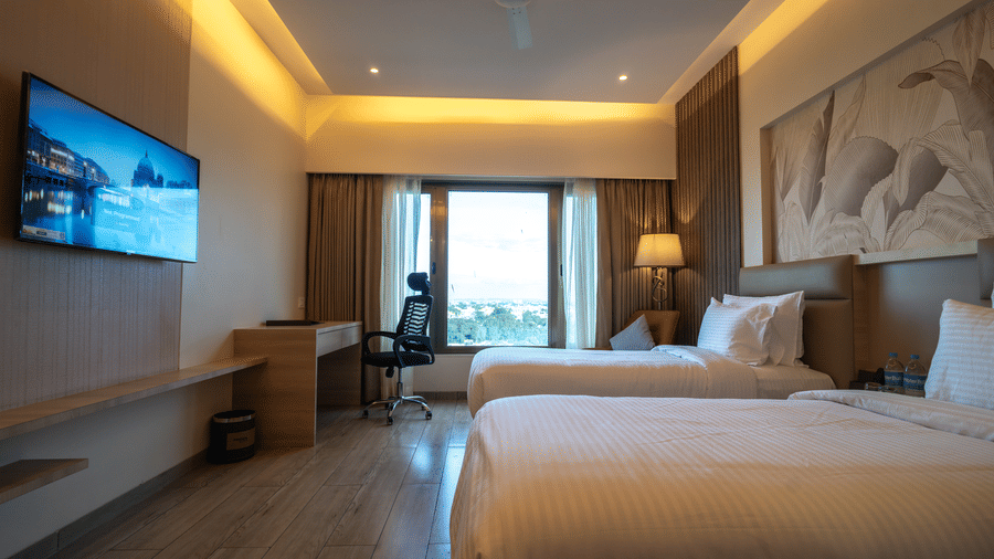 A brightly lit hotel room featuring a twin beds on the right, a wall-mounted TV on the left, and a large window behind the bed area | Pride Elite Chhatrapati Sambhajinagar