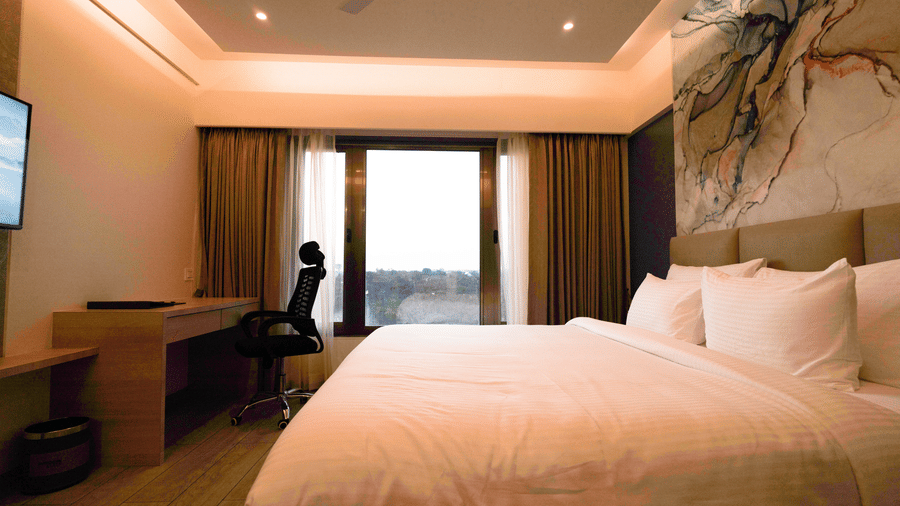 A bright hotel room featuring a double bed, a desk and chair, and a large window or balcony door | Pride Elite Chhatrapati Sambhajinagar
