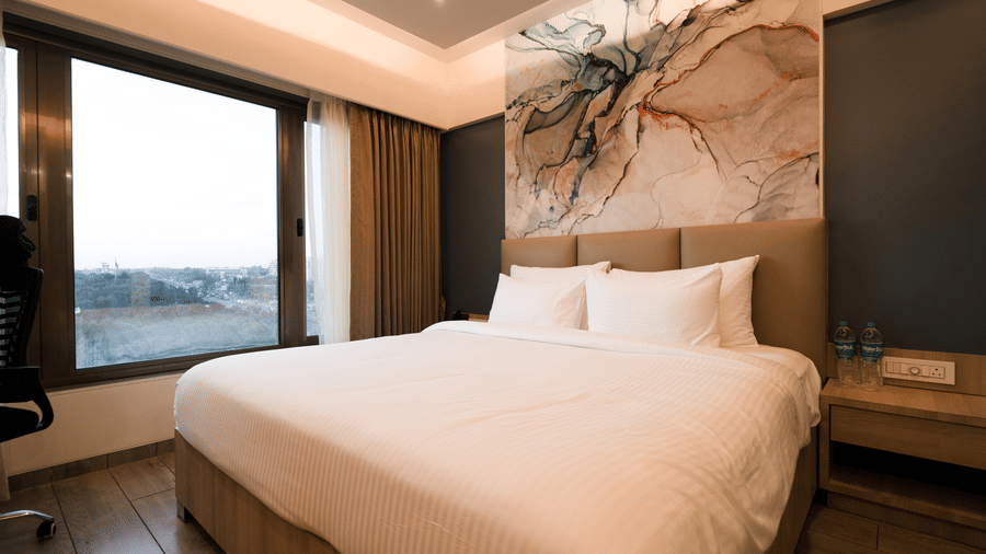 A bright hotel room featuring a double bed with white bedding, abstract art above the headboard, and a large window with outdoor views | Pride Elite Chhatrapati Sambhajinagar