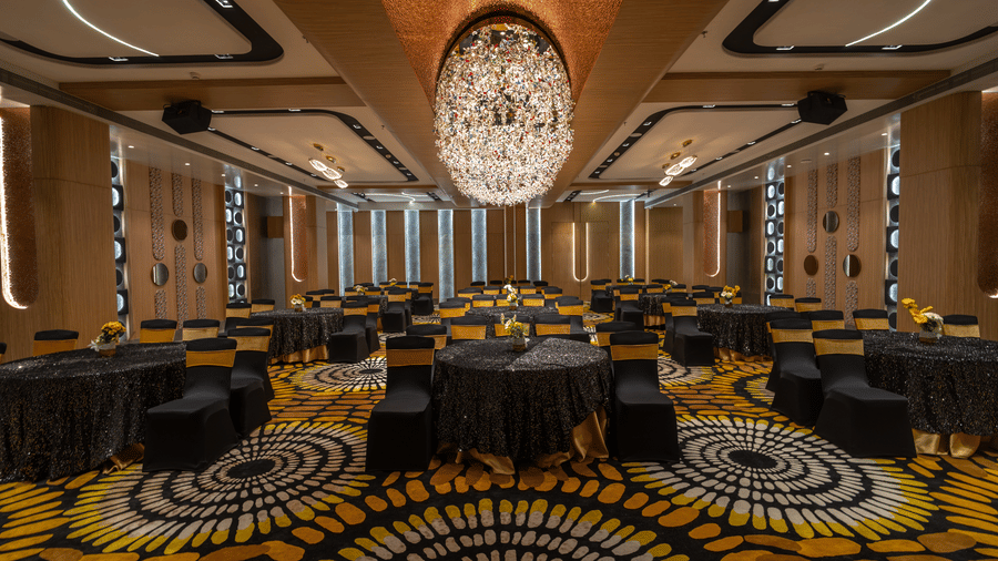 A banquet hall featuring round tables with black cloths, yellow and black patterned carpet, and elaborate linear chandelier | Pride Elite Chhatrapati Sambhajinagar