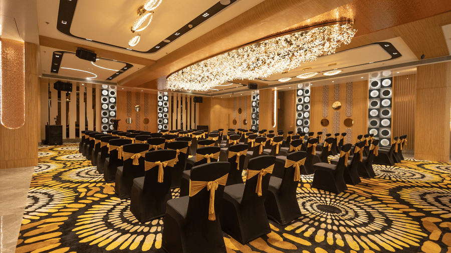 A well-lit banquet hall featuring rows of chairs with black and yellow covers and an linear chandelier | Pride Elite Chhatrapati Sambhajinagar