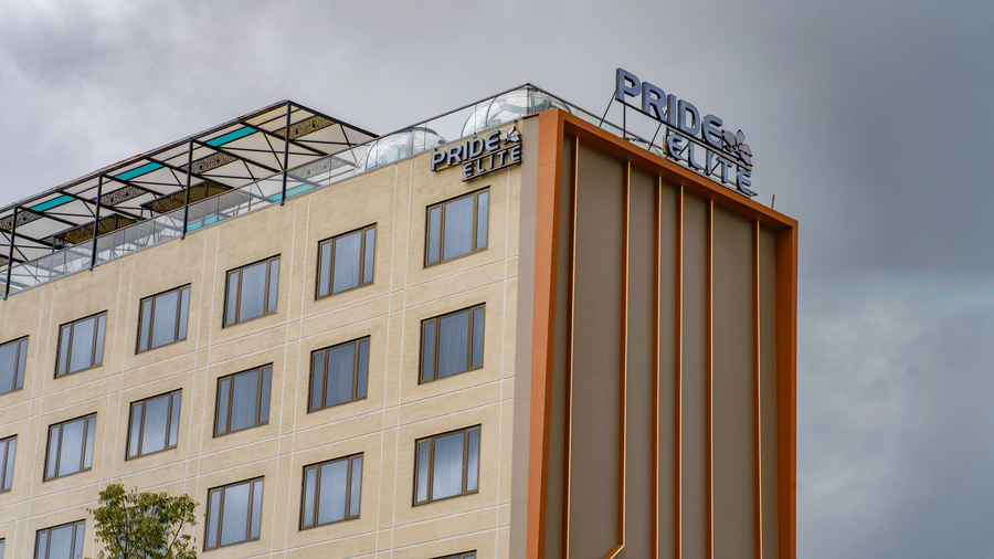 A low-angle exterior shot of a modern, multi-storey hotel building with a glass and beige facade, showing the hotel's sign on the roof against an overcast sky