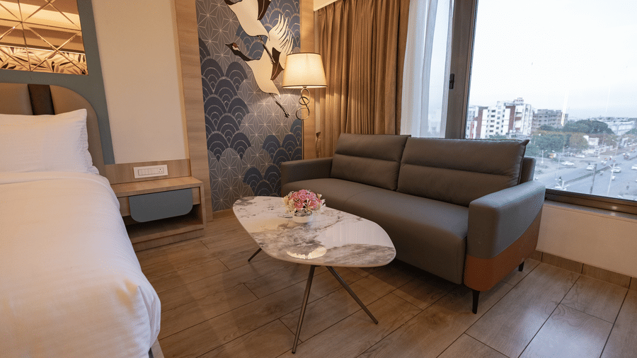 A close up view of the seating area in a hotel room, featuring a grey sofa, a small marble-topped coffee table, and a large window with city views | Pride Elite Chhatrapati Sambhajinagar