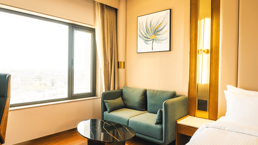 A guestroom corner showcasing a cushioned sofa, low coffee table, and framed artwork on the wall beside a double bed at Pride Plaza Indore.