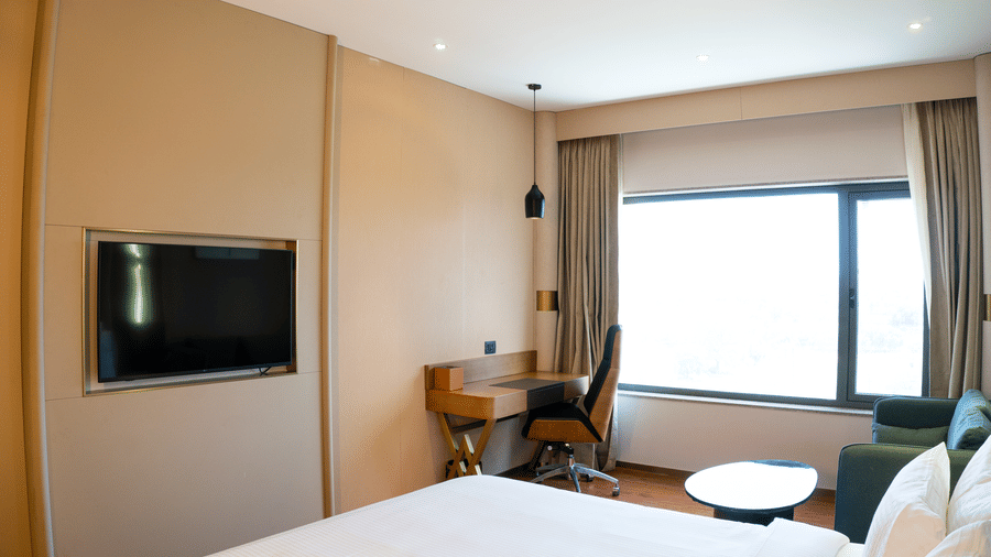 A large king size bed facing a wall mounted television screen, a wooden study table, and an armchair at Pride Plaza Indore.