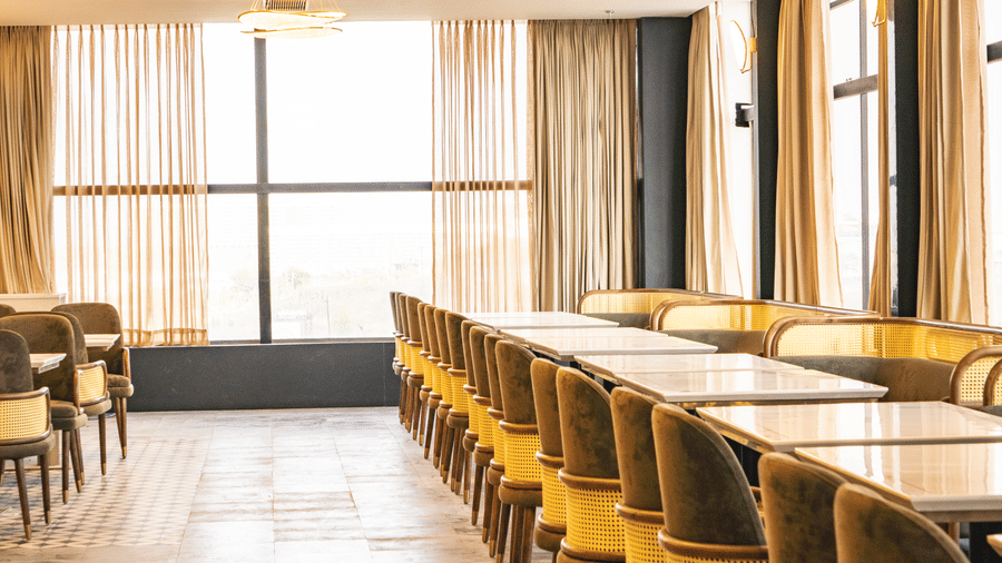 Rows of dining tables, cushioned chairs, and long curtains by the window at Pride Plaza Indore.